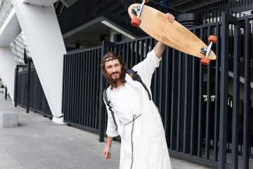 Obraz na płótnie Canvas Cheerful Jesus in robe, crown of thorns and bag showing skateboard on street
