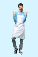 Full body of Man wearing an apron celebrating a victory and happy for having won a prize on blue background