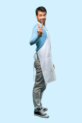 Full body of Man wearing an apron smiling and showing victory sign on blue background