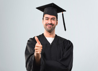 Man on his graduation day University giving a thumbs up gesture and smiling because something good has happened on grey background