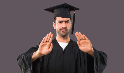 Man on his graduation day University making stop gesture with her hand for disappointed with an opinion on violet background