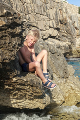 cute tanned and sunburned hair boy sitting on the edge of a steep rock on the seashore