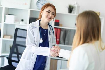 A beautiful young girl in a white coat is standing near the desk in the office and shaking hands...