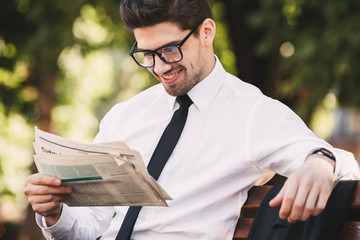 Photo of successful man in businesslike suit sitting on bench in green park, and reading newspaper...