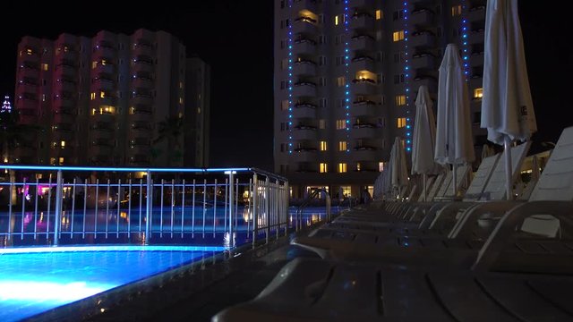 Evening, the pool area at the hotel or a multi-storey residential residence. Rows of empty chaise lounges and umbrellas. In the background of the hotel window.