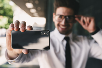 Image of businesslike man dressed in formal suit standing outside glass building, and taking selfie photo on smartphone