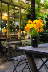Vertical yellow and orange singapore daisy flower in a brown glass jar on wooden table and wooden chair with blurred garden background