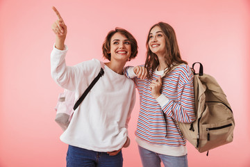 Women friends isolated over pink wall background holding backpacks pointing.