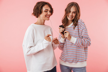 Happy women friends isolated over pink wall background listening music with earphones.