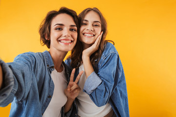 Excited young women friends isolated over yellow wall background take a selfie by camera.