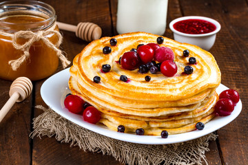 Pancakes with fresh berries of blueberries and cherries, raspberry jam, milk and honey on a wooden background. Delicious, healthy breakfast