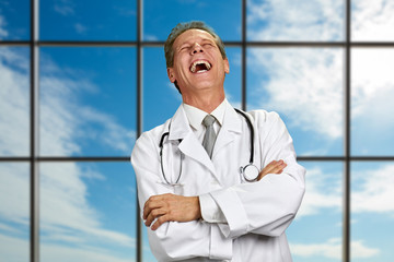 Mature doctor is laughing on sky background. Joyful medical specialist crossed arms at hospital...