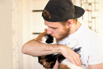 Close-up of beard man in icelandic sweater who is holding and kissing his cute purring Devon Rex cat. Muzzle of a cat and a man's face. Love cats and humans. Relationship, weasel.