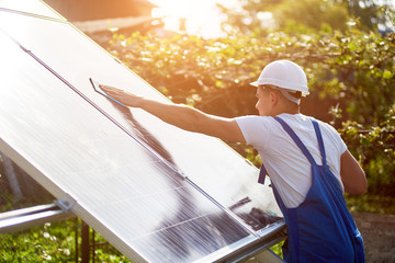 Profile of worker in white helmet and blue overalls wiping clean big square shiny solar panel on...