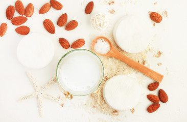 Almond milk and rice water for holistic skin care therapy. Top view white table background with jars, nuts, cereal. Cleansing foam in spoon. Beige and white light colors 