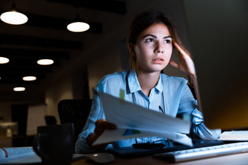 Woman graphic designer using pc computer working with documents at night in office.