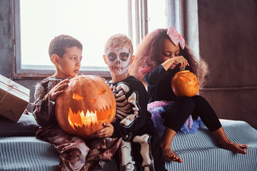 Group of multiracial kids during Halloween party holds pumpkins while sitting on bed in an old...