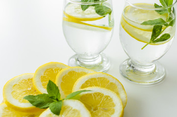 Two cold drinks in transparent glasses next to plate full of lemon slices with fresh mint leaves on them.