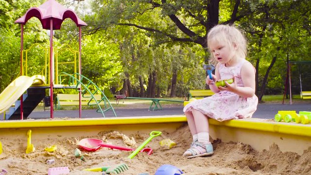 Low angle view. A little girl sitting in a sandbox is picking up sand with a scoop in a mold.