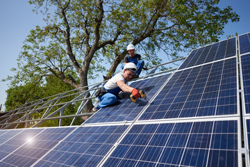 Two workers on tall steel platform installing heavy solar photo voltaic panel using screwdriver on green tree and blue sky background. Solar panel system installation, professionalism concept.