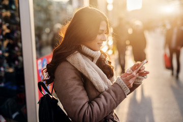 Cute young girl in winter coat standing in street . Holding telephone and looking away. Sunny winter day.