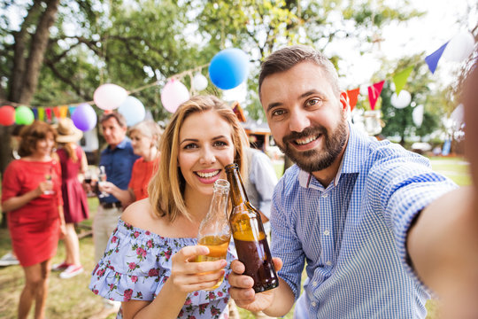 Young couple taking selfie at a party outside in the backyard, clinking bottles.