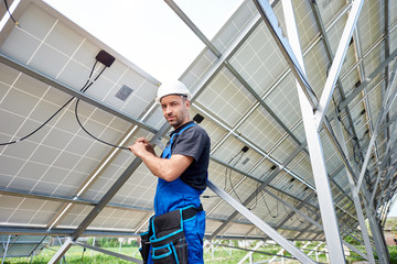 Young engineer technician making electrical wiring standing inside high exterior solar panel photo voltaic system, looking to the camera. Eco friendly cheap electricity generation concept.