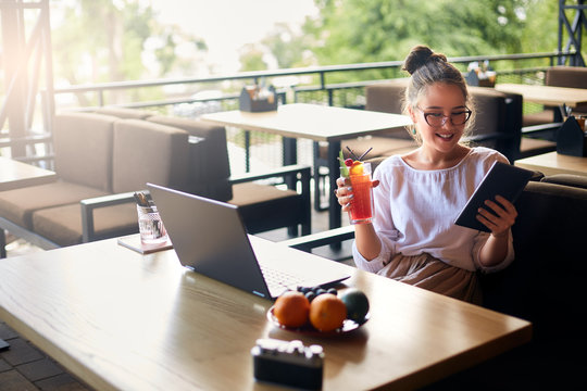 Smiling Mixed Race Woman With Cocktail In Hand Works With Laptop. Businesswoman In Glasses Drinks Juice For Body Hydration While Working. Attractive Designer Quench Thirst. Healthy Lifestyle Theme.