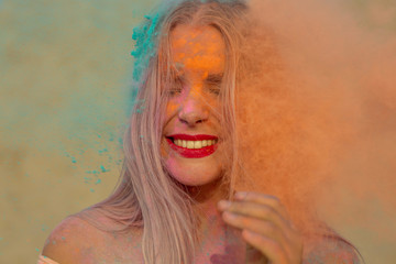 Closeup shot of adorable blonde model with red lips playing with exploding orange and blue dry paint Holi at the desert