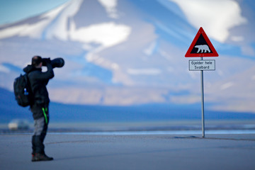 Photographe avec grand objectif et panneau de signalisation routière avec ours polaire. &quot Gjelder Hele Svalbard&quot  signifie &quot sur tout le Svalbard (attention aux ours polaires)&quot . Homme sur la route avec la montagne enneigée, Svalbard.