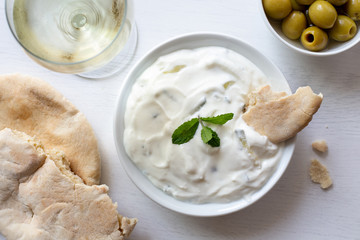 Tzatziki in white ceramic bowl with mint leaf garnish and a piece of pita bread next to pita bread, olives and a glass of white wine from above.