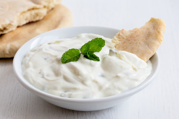 Tzatziki in white ceramic bowl with mint leaf garnish and a piece of pita bread next to pita bread.