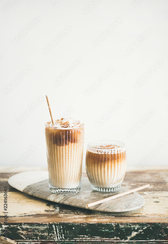 Poster iced coffee in tall glasses with milk and straws on board over wooden table, white wall at backgroun