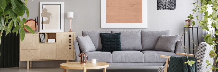 Real photo of light grey living room interior with wooden cupboard, two posters, grey couch with pillows and round coffee table with lantern and cup