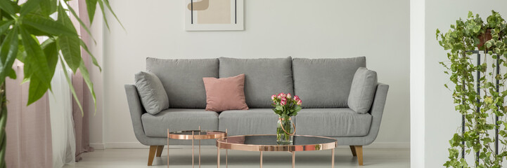 Real photo of grey settee with dirty pink pillow standing in white living room interior with fresh plant and roses in glass vase placed on metal table