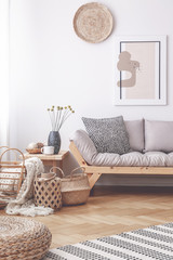 Baskets and pouf on wooden floor in living room interior with poster above grey sofa. Real photo