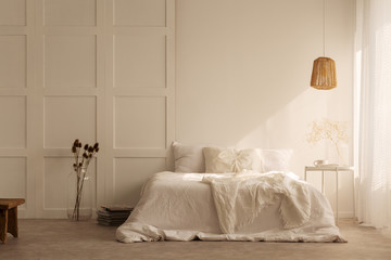 Lamp above white bed with pillows in minimal bedroom interior with plants and stool. Real photo - obrazy, fototapety, plakaty