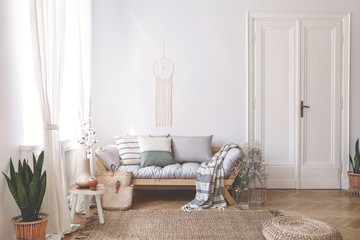 Blanket and pillows on wooden sofa in white loft interior with pouf and plant on carpet. Real photo