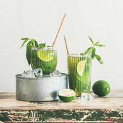 Green matcha ice-tea with lime and mint in glasses over rustic cupboard, white wall at background,...