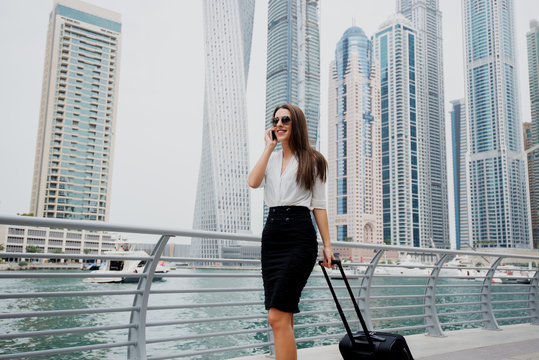 Busy young modern business woman in suit pulling a suitcase in a Dubai Marine. Walking to her office and talking on a telephone..