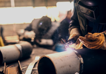 Professional welders in protective uniform and mask welding metal pipe in workshop.