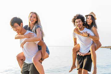 Group of friends walking along the beach, with men giving piggyback ride to girlfriends. Happy...