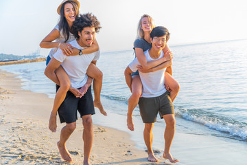 Group of friends walking along the beach, with men giving piggyback ride to girlfriends. Happy...