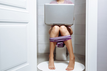 Young woman is using laptop while seated on toilet in morning.