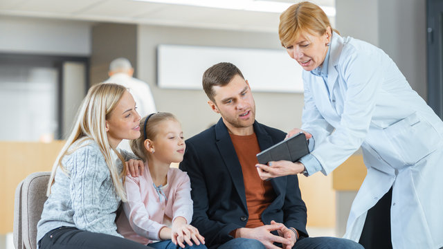 In The Hospital Lobby Female Doctor Shows Information On The Tablet Computer To The Young Family (Father, Mother And Little Daughter). They All Serious And Have Bad News. Modern Medical Facility.