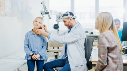 Fototapeta premium Matka z córeczką odwiedza lekarza. Przyjazny pediatra laryngolog sprawdza uszy małej uśmiechniętej dziewczynki. Badanie zdrowia dziecka przy użyciu specjalnego sprzętu latarki.