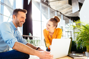 Two cheerful business partners chatting during meeting in restaurant and looking at laptop