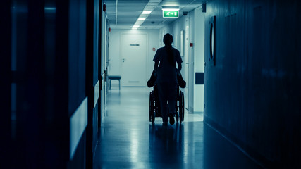 Silhouette of the Female Nurse Moving Patient in the Wheelchair in the End of Hospital Corridor....