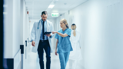 Female Surgeon and Doctor Walk Through Hospital Hallway, They Consult Digital Tablet Computer while...