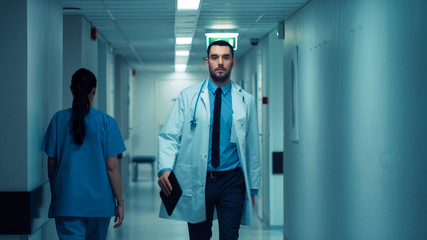 Determined Handsome Doctor Wearing White Coat with Stethoscope Walks Through Hospital Hallway....